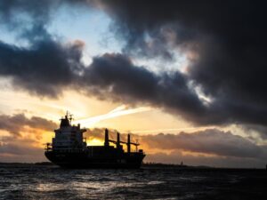 A large cargo ship sailing across the water at sunset beneath dramatic dark clouds