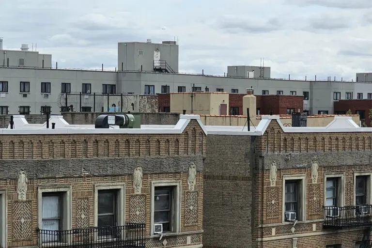 A brick residential building rooftop with parapet walls, windows, air-conditioning units, and utility structures, with additional buildings visible behind