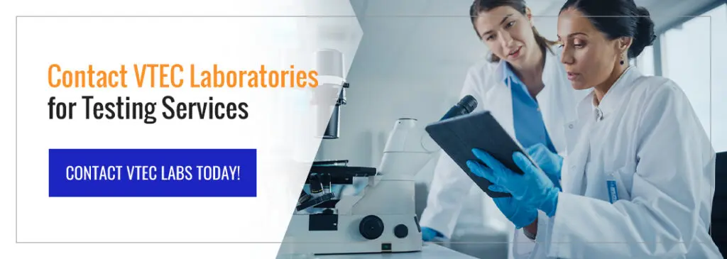 Two laboratory technicians wearing protective gloves review test data on a tablet next to a microscope in a testing laboratory.